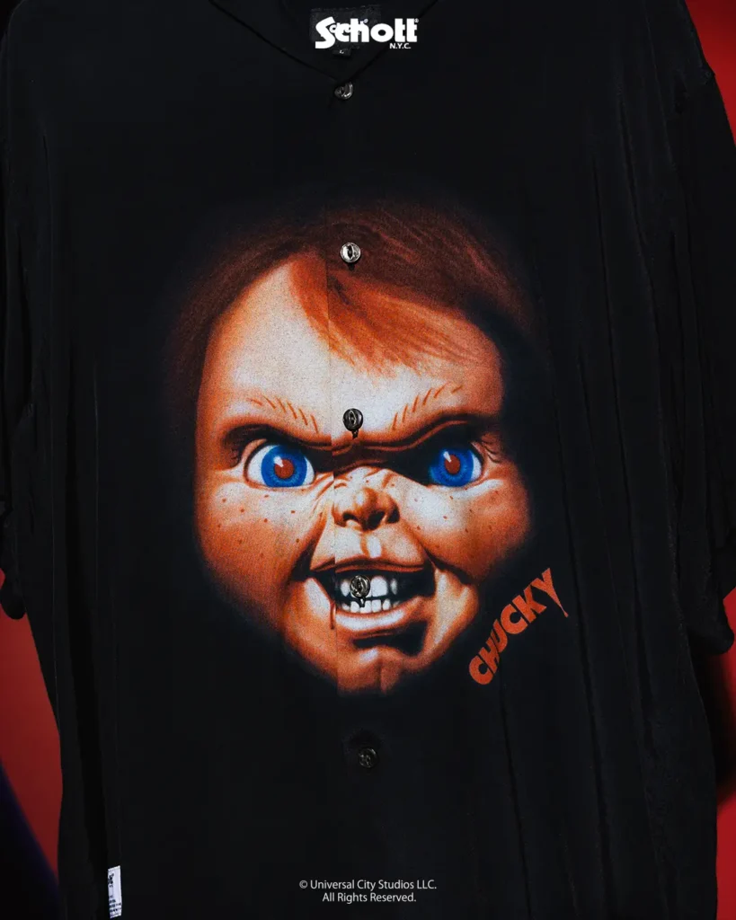 Close-up of a black Schott NYC camp-collar shirt featuring a large vintage-style graphic of Chucky’s menacing face across the front, accented by vivid blue eyes and orange lettering against a dark backdrop