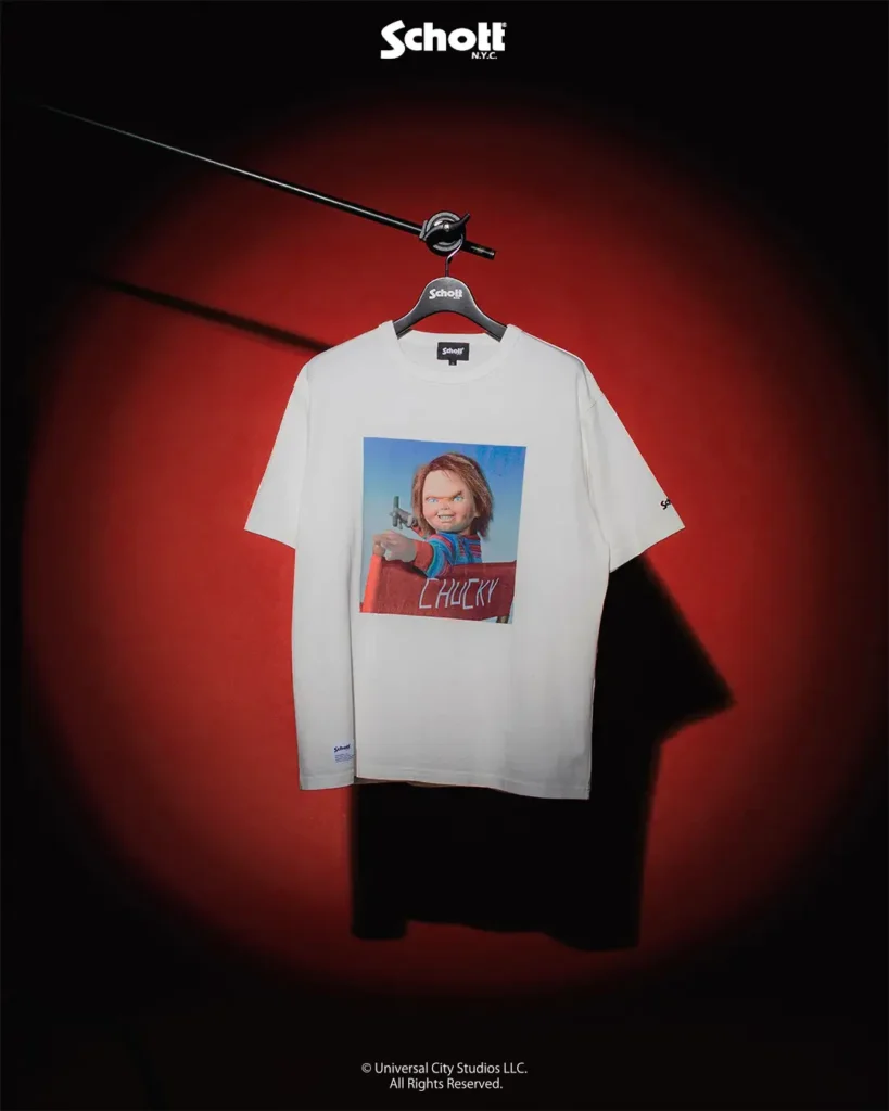White graphic T-shirt from the Schott NYC x Chucky collection featuring a colorful image of Chucky pointing a handgun, displayed against a red-and-black theatrical background