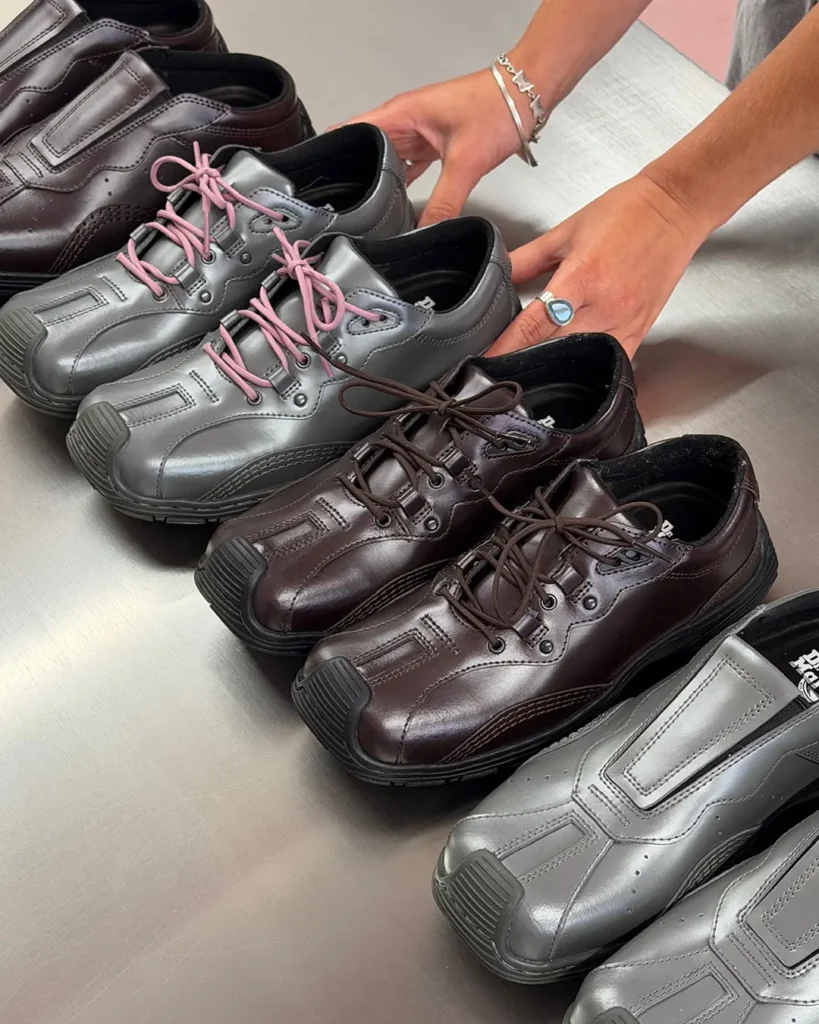 A selection of The Aurix leather shoes is displayed on a brushed metal table in dark brown and metallic silver colorways. Both lace-up and slip-on styles are shown, highlighting interchangeable lace colors, smooth leather uppers, reinforced rubber toe bumpers, and durable black soles as hands arrange the footwear