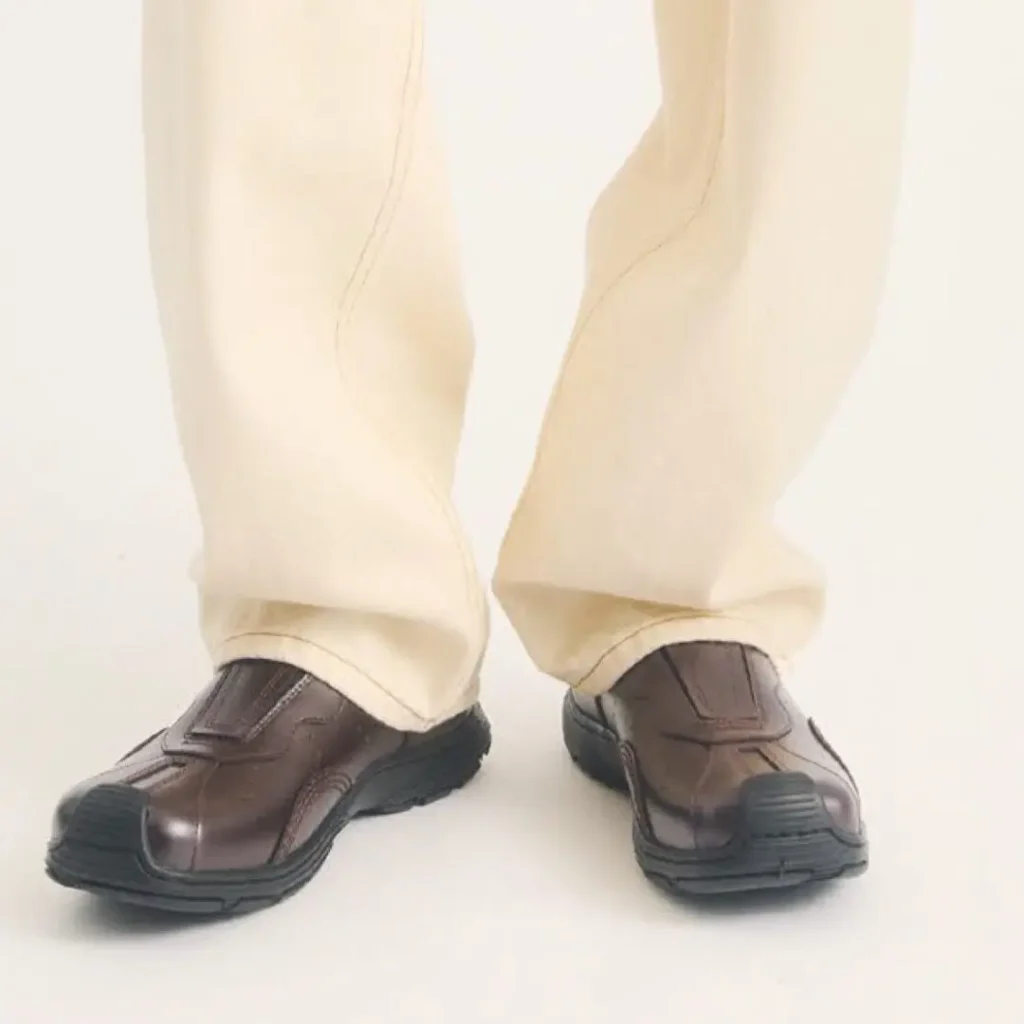 Dark brown leather slip-on versions of The Aurix are paired with relaxed cream trousers in a bright studio environment. The shoes feature a protective leather instep cover, reinforced rubber toe, cushioned black sole, and polished leather upper that balances classic styling with functional comfort