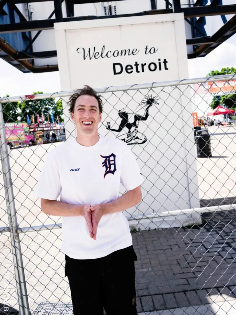 Welcome to Detroit – A smiling person stands in front of a "Welcome to Detroit" sign wearing a white Palace x Detroit T-shirt with a navy Detroit “D” logo and Palace wordmark