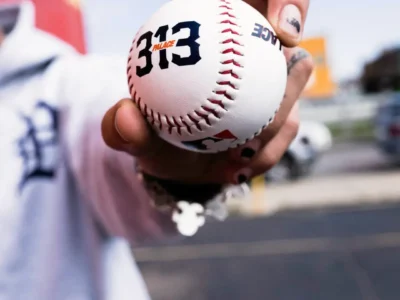 Close-up of baseball – A hand with black-painted fingernails grips a white baseball featuring Palace branding, the number “313,” and an MLB logo, with a person in a Detroit-inspired jersey blurred in the background