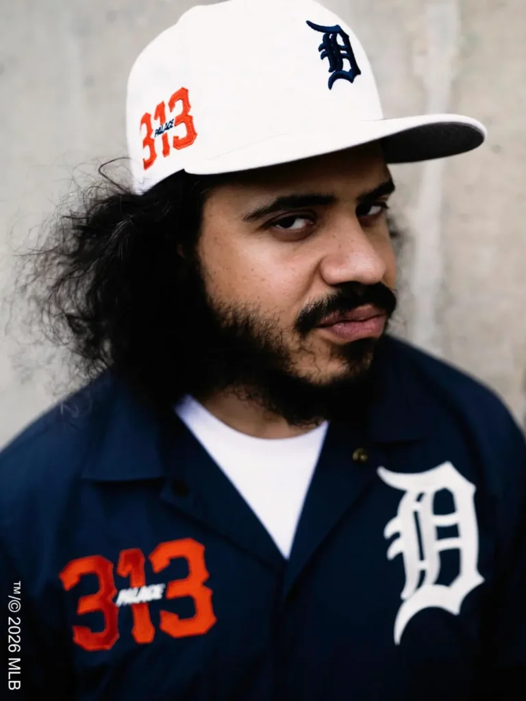 Portrait in Palace x Detroit gear – A person with long curly hair and a beard wears a white Palace x Detroit baseball cap and navy overshirt with oversized “D” and “313” graphics, posing against a concrete wall