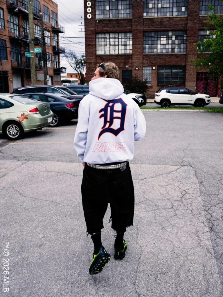Gray hoodie from behind – A person walks through an industrial Detroit street wearing a light gray Palace hoodie with a large Detroit “D” logo and Palace branding across the back
