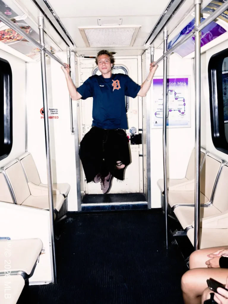 Subway pose – A person stands inside a subway car holding the overhead rails while wearing a navy Palace x Detroit T-shirt with a large orange Detroit “D” logo on the front