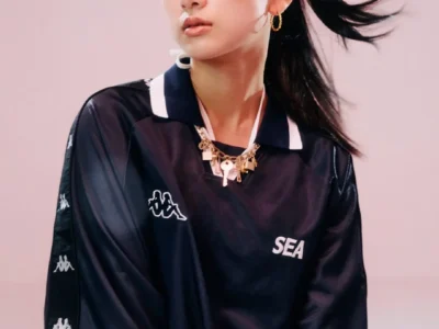 Fashion portrait of a woman wearing a black Kappa x Wind And Sea football-inspired jersey with gold jewelry and a high ponytail, posing against a soft pink studio backdrop