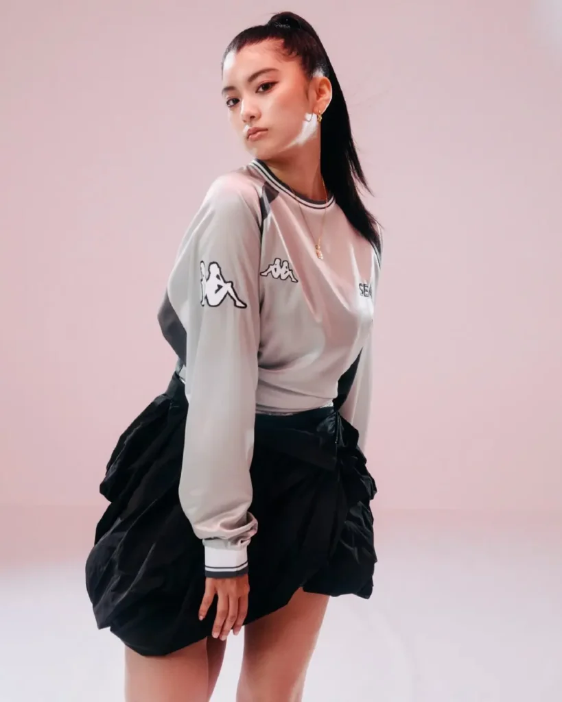 Full-body fashion portrait featuring a silver-gray long-sleeve Kappa top and voluminous black skirt, styled against a clean pastel pink background