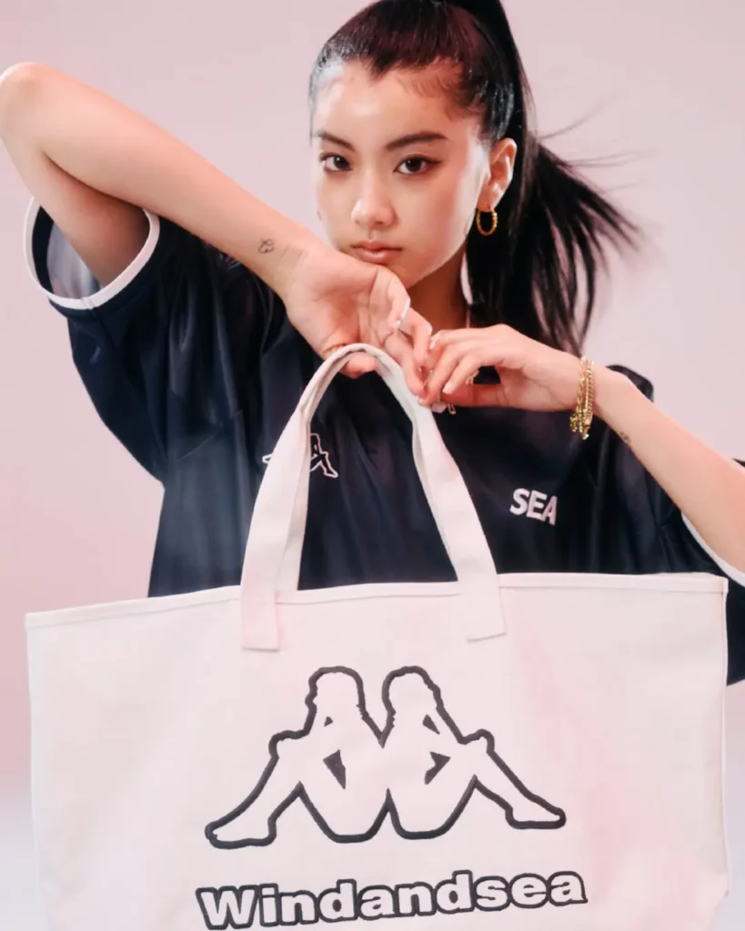 Close-up portrait of a woman holding a large Wind And Sea tote bag, wearing a black jersey top with gold jewelry and a sleek high ponytail