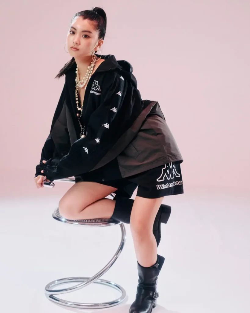 Editorial fashion shot of a woman seated on a chrome stool wearing a black Wind And Sea jacket, logo shorts, layered pearls, and black boot