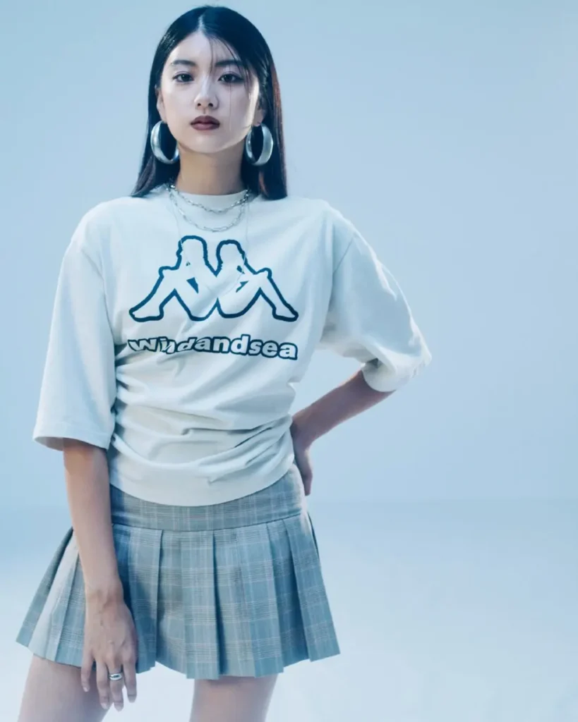 Woman standing in a pleated plaid mini skirt, oversized Wind And Sea logo tee, and statement earrings against a minimalist blue backdrop