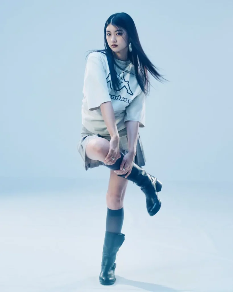 Full-body fashion portrait of a woman wearing a Wind And Sea graphic T-shirt, plaid skirt, knee-high socks, and black boots in a blue-toned studio