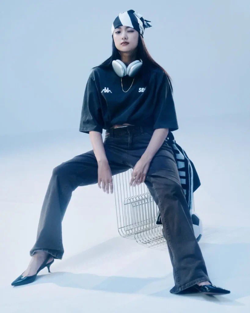 Woman seated on a chrome wire chair wearing a black oversized Wind And Sea tee, dark denim, pointed heels, and silver headphones, styled in a cool blue-toned studio set