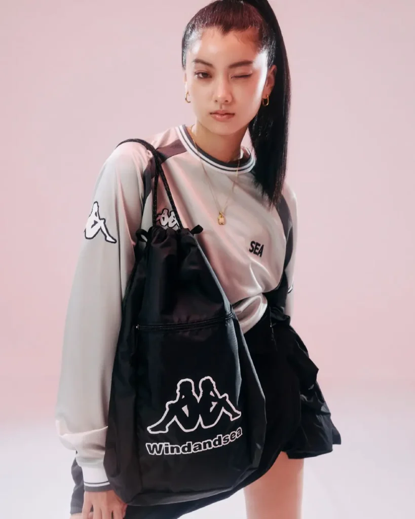 Woman winking while carrying a black Wind And Sea drawstring bag, styled in a silver-gray Kappa jersey top and black skirt against a pink backdrop