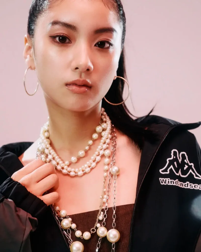 Close-up editorial portrait featuring layered pearl necklaces, hoop earrings, and a black Wind And Sea jacket against a softly lit pink background