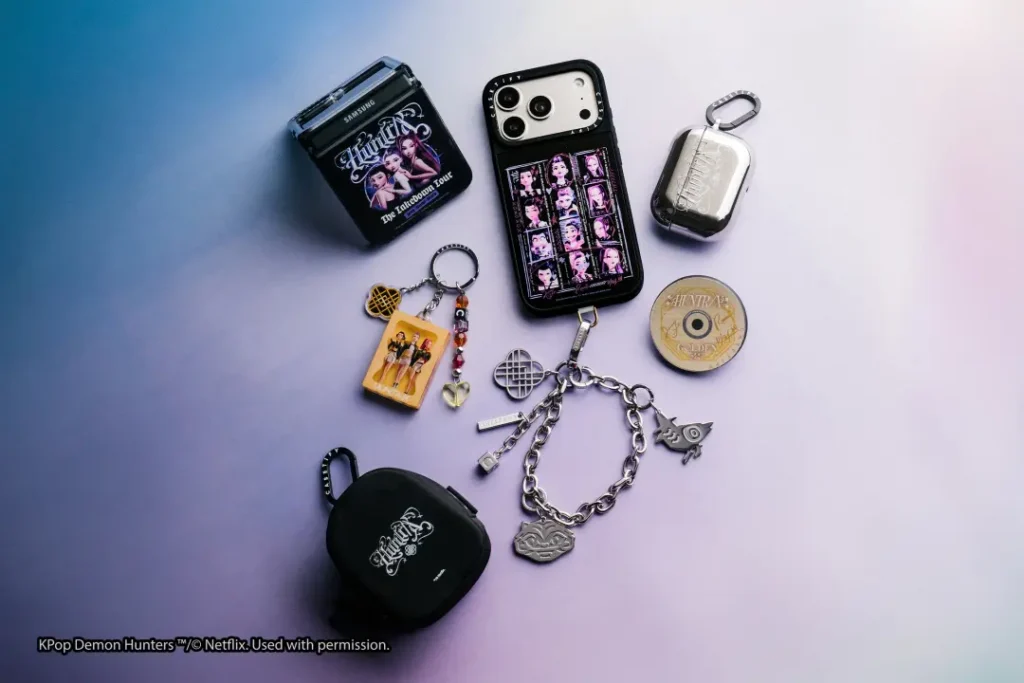 Flat lay of KPop Demon Hunters-themed CASETiFY accessories including a smartphone case, Galaxy Z Flip case, AirPods case, keychains, phone charms, wrist chain, and collectible disc arranged on a soft gradient background