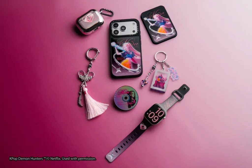 Pink-themed CASETiFY KPop Demon Hunters collection featuring a Mira character phone case, matching magnetic wallet, AirPods case, Apple Watch band, keychains, tassel charm, and decorative collectible disc on a pink gradient surface