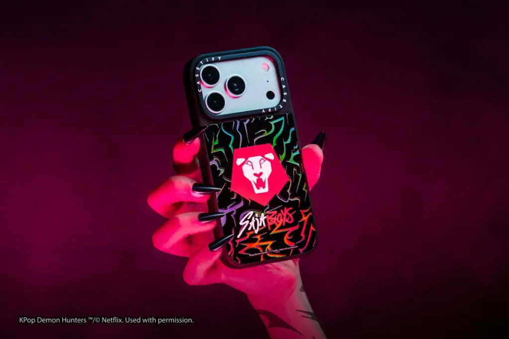 Close-up of a CASETiFY smartphone case inspired by the Saja Boys from KPop Demon Hunters, featuring bold graffiti graphics and a red emblem, held dramatically under vivid magenta studio light