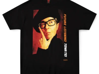 Black short-sleeve graphic T-shirt featuring a large vintage-style portrait of Towa Tei wearing thick black glasses and a dark hat. Vertical orange and white text reading “FUTURE LISTENING! TOWA TEI” runs alongside the artwork, with a small stylized logo near the lower right of the print. The shirt is displayed flat against a white background