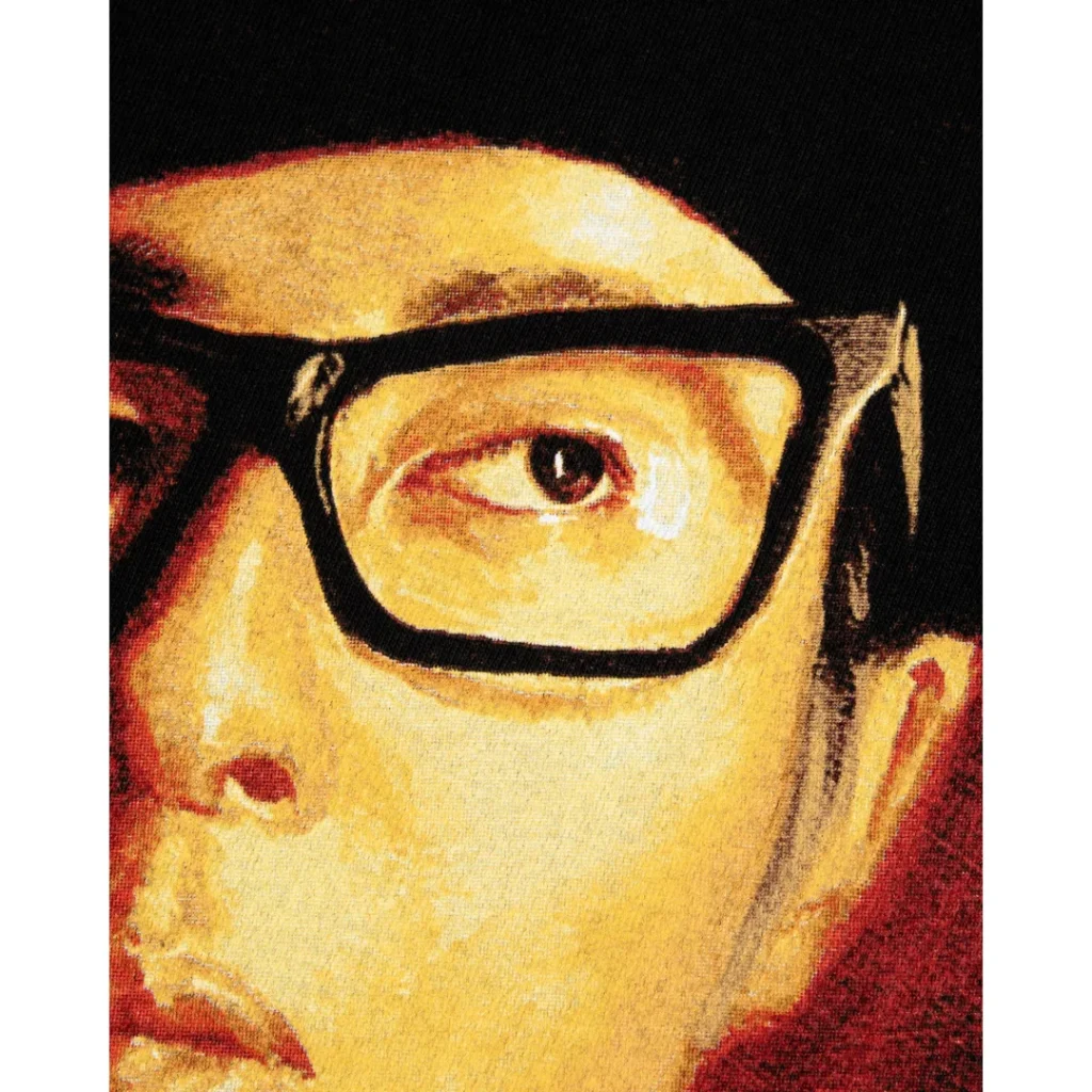 Detailed close-up of the printed portrait highlighting Towa Tei’s face, oversized black-framed glasses, and eye. The textured artwork uses warm gold, brown, and red tones, emphasizing the vintage, painterly print quality on the black cotton fabric