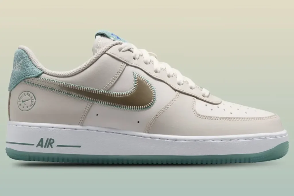 Nike Air Force 1 Low in a clean sail leather construction featuring a reflective metallic silver Swoosh outlined with aqua stitching. Soft terry-cloth heel tabs in pastel blue add texture, while translucent icy-blue outsoles provide a fresh contrast. Subtle Philly-inspired branding appears near the heel, with matching aqua accents on the midsole “AIR” logo