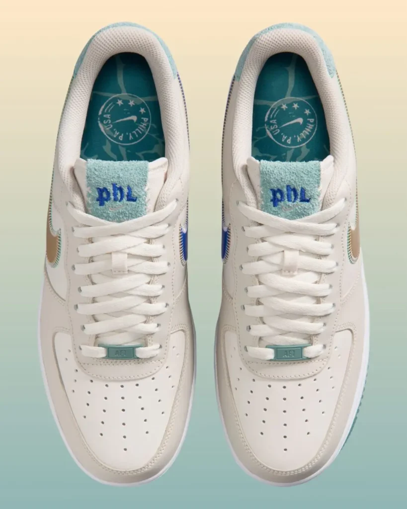 Overhead view of the Nike Air Force 1 Low showcasing perforated sail leather toe boxes, white flat laces, and pastel aqua lace dubraes. Plush terry-cloth tongues feature embroidered blue “pbl” lettering, while teal insoles display custom Philadelphia-themed graphics and branding detail