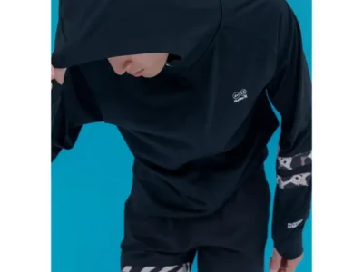Model wearing a black hooded performance jacket from the Hurley x uniform experiment collaboration, featuring a concealed face design and graphic patterned detailing on the sleeve against a bright blue backdrop