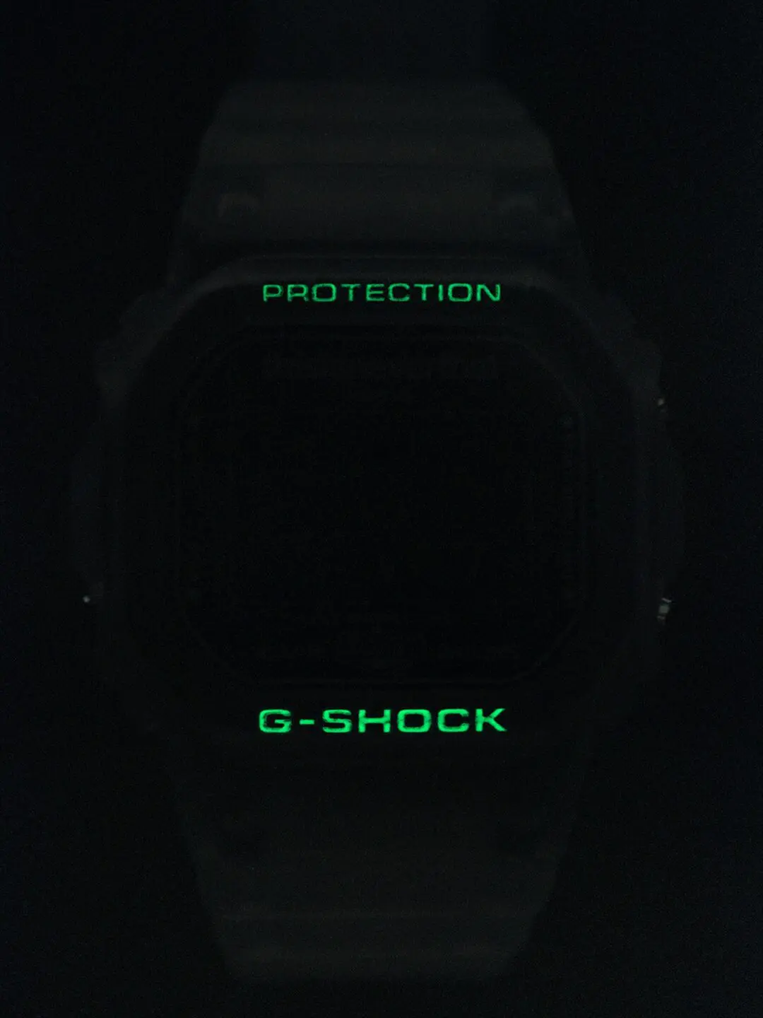 Glow-in-the-dark view of the thisisneverthat x G-SHOCK digital watch. In near-total darkness, the translucent case fades into the background while the “PROTECTION” and “G-SHOCK” markings emit a bright green glow, emphasizing the watch’s luminous detailing and rugged sil
