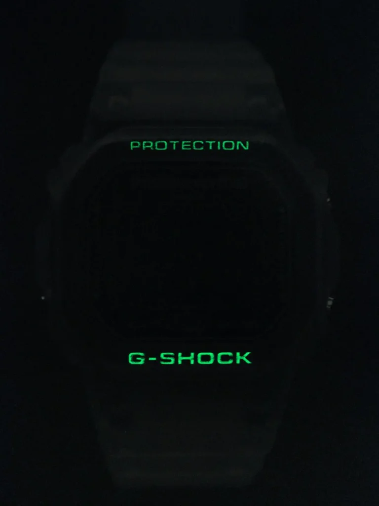 thisisneverthat x G-SHOCK DW-6900TNT24-7DF in total darkness with signature green EL backlight illuminating "PROTECTION" above the dial and bold "G-SHOCK" below — the fourth collaboration’s subtle illuminated detail on the milky ivory Triple Graph model