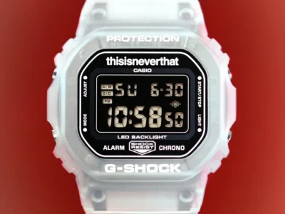 Front-facing product shot of the thisisneverthat x G-SHOCK watch against a vivid red backdrop. The translucent frosted resin case and strap frame a black digital display, while collaborative branding appears above the screen alongside classic G-SHOCK text and functionality mark