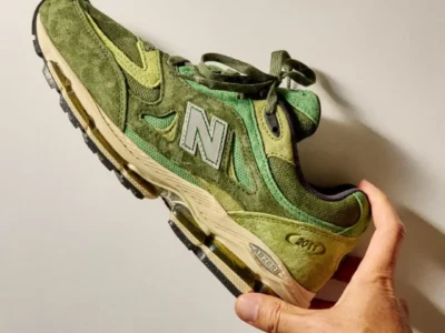 A hand holds a New Balance 2011 sneaker in a rich leaf-green colorway against a neutral wall. The shoe features layered suede and mesh panels in varying shades of olive, moss, and lime green, accented by a silver reflective “N” logo. An aged cream midsole and visible ABZORB cushioning add a vintage-inspired finish, while warm natural light casts a dramatic sil behind the shoe