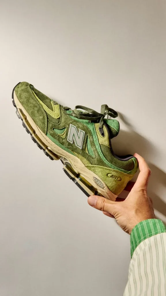 A hand holds a New Balance 2011 sneaker in a rich leaf-green colorway against a neutral wall. The shoe features layered suede and mesh panels in varying shades of olive, moss, and lime green, accented by a silver reflective “N” logo. An aged cream midsole and visible ABZORB cushioning add a vintage-inspired finish, while warm natural light casts a dramatic sil behind the shoe