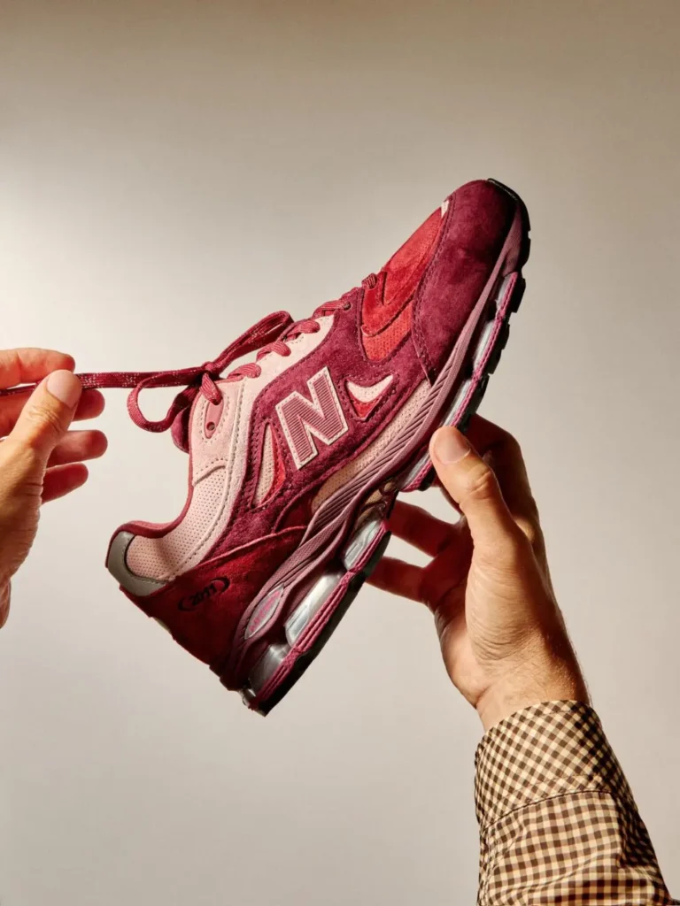 Close-up studio photograph of a burgundy and pink New Balance 2010 shoe held in both hands. Rich suede overlays, perforated panels, reflective accents, and view ABZORB cushion bare highlighted by warm lighting, emphasizing the shoe’s premium craft and layered tincture palette