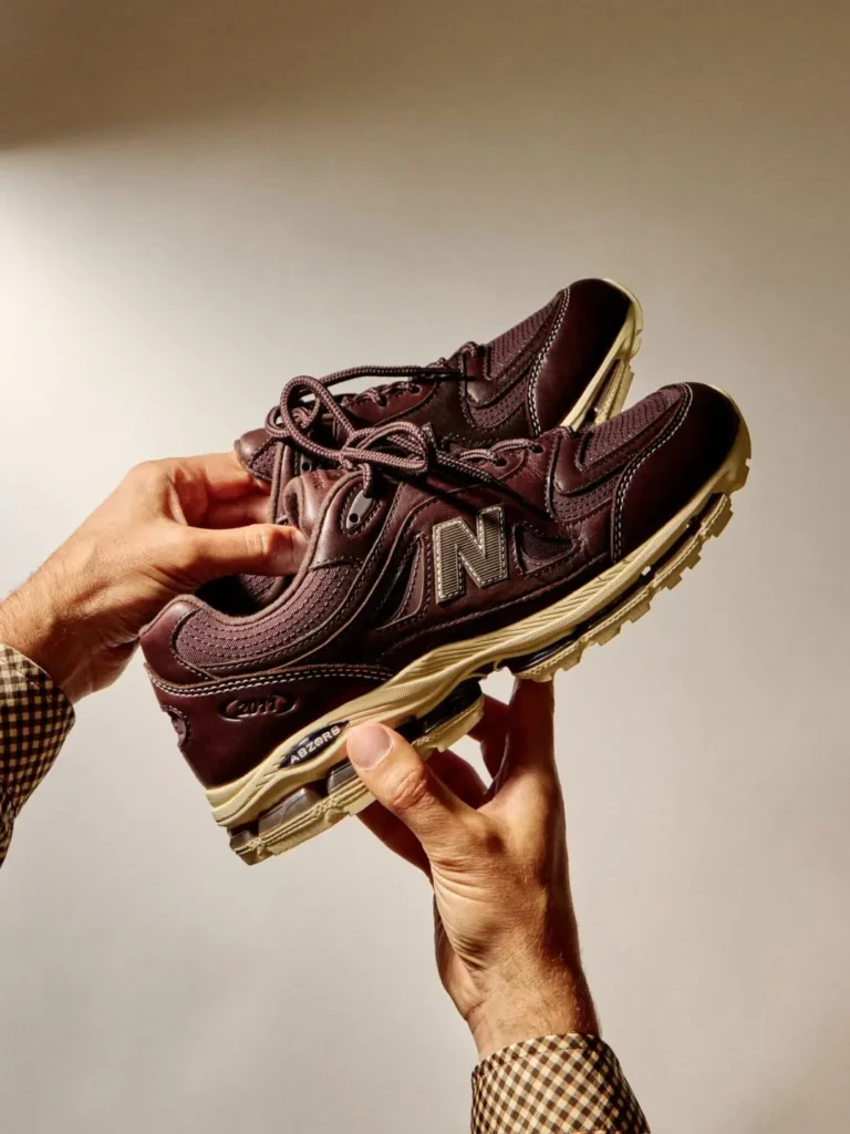 Close-up studio photograph of a dark brown New Balance 2010 shoe held in both hands. Premium leather and mesh materials, contrast stitching, metallic “N” branding, and a cream ABZORB sole unit are showcased through dramatic warm lighting and a min backdrop