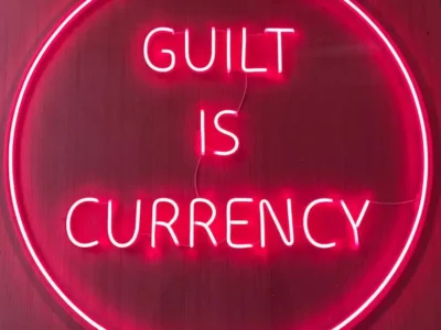 Neon pink artwork featuring the phrase “GUILT IS CURRENCY” glowing inside a circular light installation against a dark background, creating a bold contemporary statement on value, emotion, and social exchange