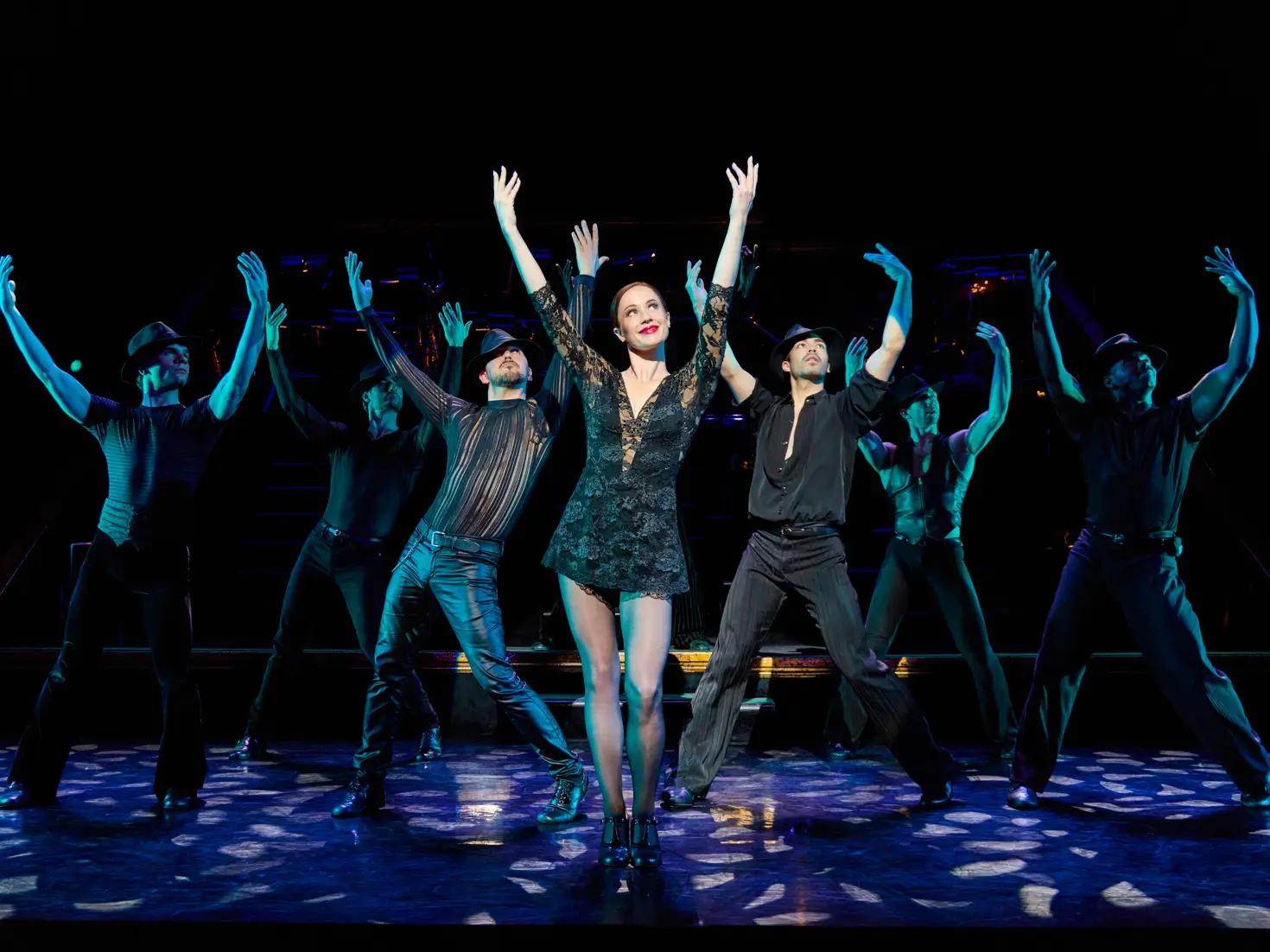 A theatrical stage performance features a lead dancer in a black lace dress standing center stage with arms raised, surrounded by dancers in dark costumes and hats executing synchronized choreography under dramatic blue and spotlight lighting