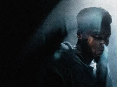 Grainy cinematic portrait of a man standing in deep sil beneath a beam of cool blue light. The low-resolution, film-inspired image features heavy texture, soft focus, and atmospheric haze, creating a moody and introspective visual reminiscent of vintage analog photo