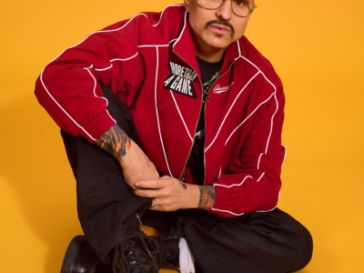 Portrait of a person seated against a vibrant yellow backdrop, wearing a red track jacket with white piping, black pants, and black lace-up boots. Gold-framed glasses, tattoos, and a trimmed mustache complete the bold look