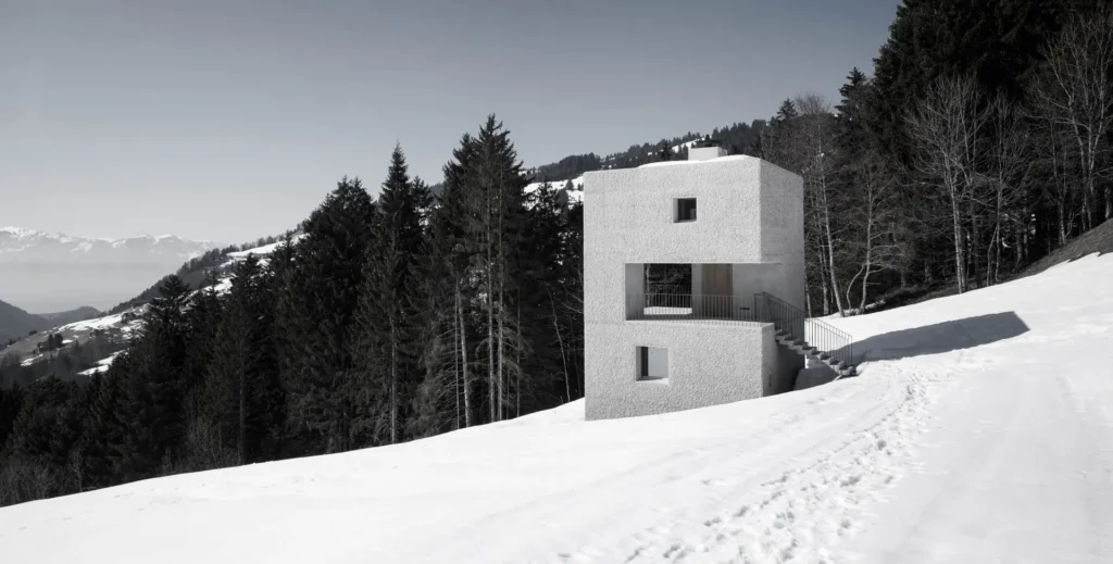 Wide alpine landscape featuring scattered snowfields, dark evergreen trees, rustic wooden buildings, and the modern cabin perched in the distance on a hillside