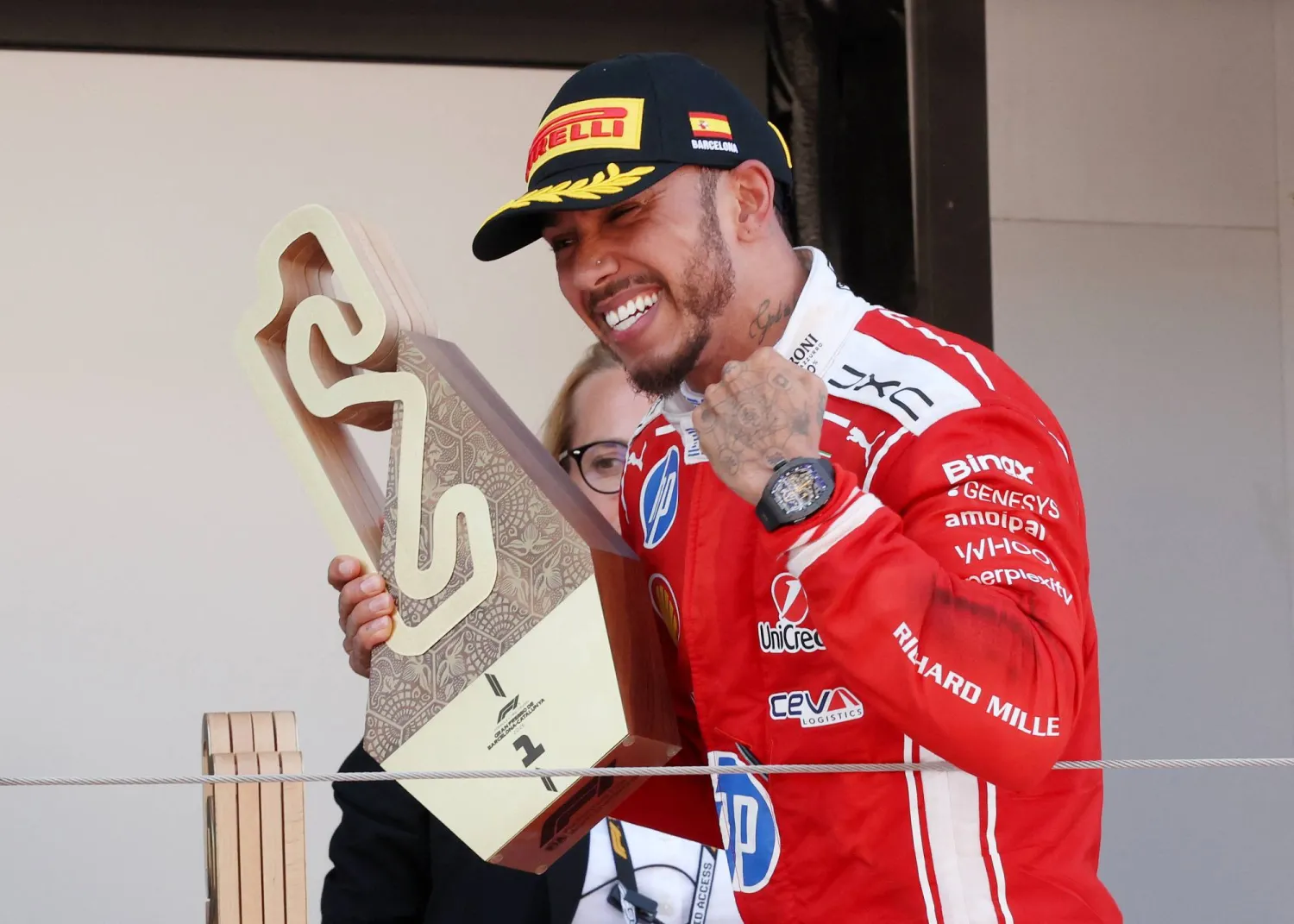 A Formula 1 driver in a red racing suit celebrates on the podium while holding a sculptural race trophy. Wearing a black cap with yellow detailing, he smiles broadly and raises a clenched fist in triumph. Sponsor logos cover the suit, and a luxury sports watch is visible on his wrist as he stands against a neutral paddock backdrop