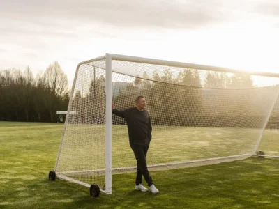 Phil McCartney stands beside a full-size football goal on a sunlit grass field, reflecting his lifelong connection to sport and athlete-driven innovation at Nike