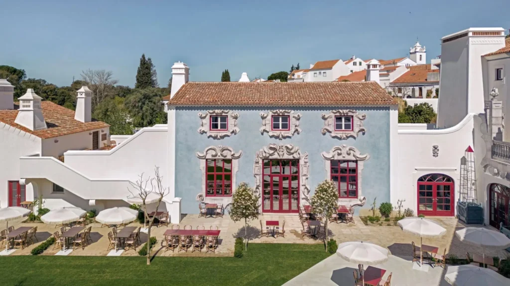 Exterior courtyard of Vermelho Hotel in Melides, Portugal, featuring a powder-blue façade, ornate Baroque-inspired window frames, red accents, and landscaped outdoor dining space