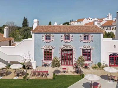 Exterior courtyard of Vermelho Hotel in Melides, Portugal, featuring a powder-blue façade, ornate Baroque-inspired window frames, red accents, and landscaped outdoor dining space
