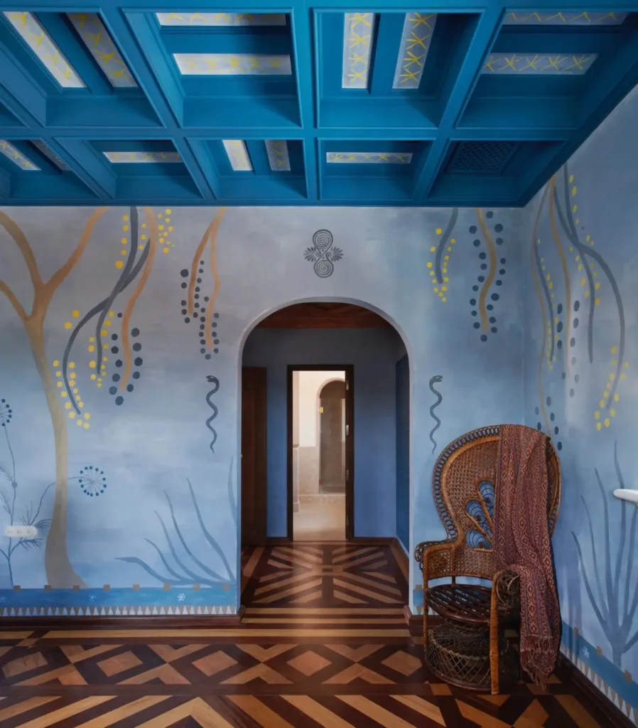 Art-filled guest room at Vermelho Hotel featuring hand-painted blue murals, a dramatic coffered ceiling, geometric wood flooring, and a woven peacock chair