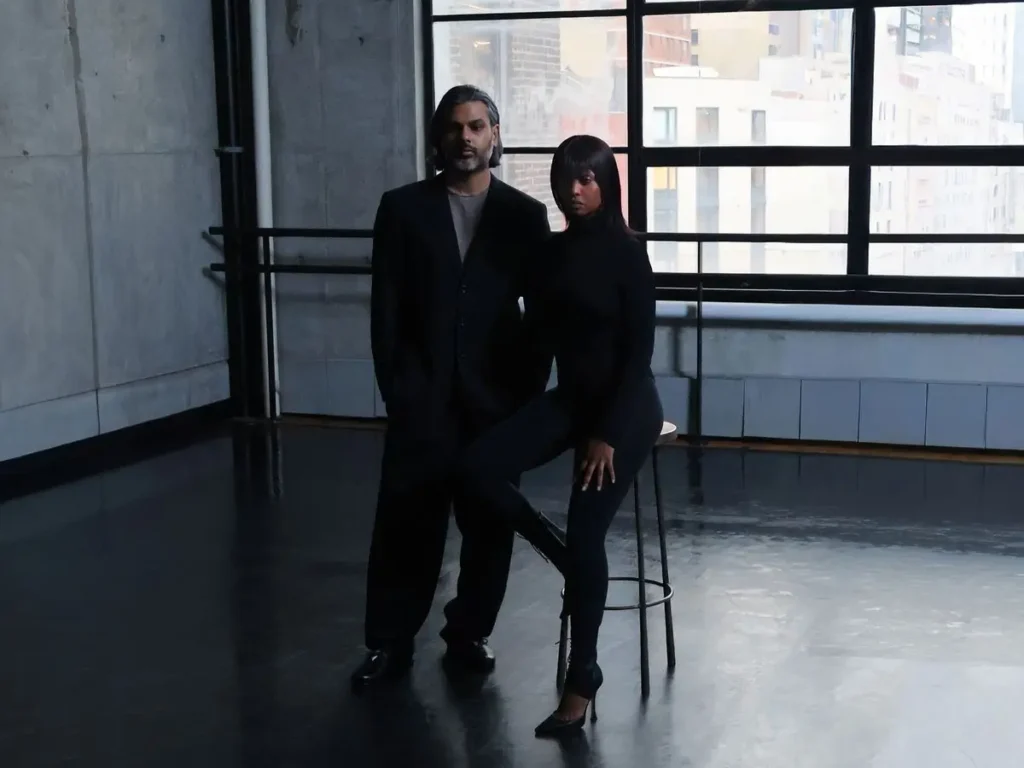 Two fashion creatives pose in a spacious industrial loft with polished concrete walls and large grid windows overlooking a cityscape. Dressed in monochromatic black tailoring and form-fitting silhouettes, they stand and sit beside a tall stool, creating a minimalist, editorial composition defined by dramatic natural light, reflections on the dark floor, and a moody contemporary atmosphere