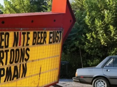 Vintage drive-in theater marquee advertising a video night and beer bust event, with a parked gray sedan beside the sign and lush green trees in the background