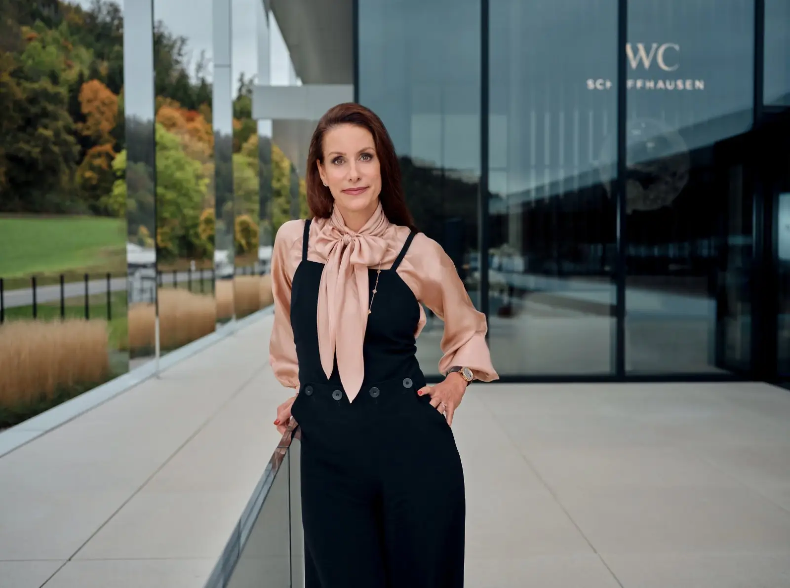Executive portrait of a woman standing outside IWC Schaffhausen headquarters, wearing a blush silk bow blouse layered under a tailored black jumpsuit. The modern glass-and-steel architecture and autumn landscape in the background reinforce a refined -business setting