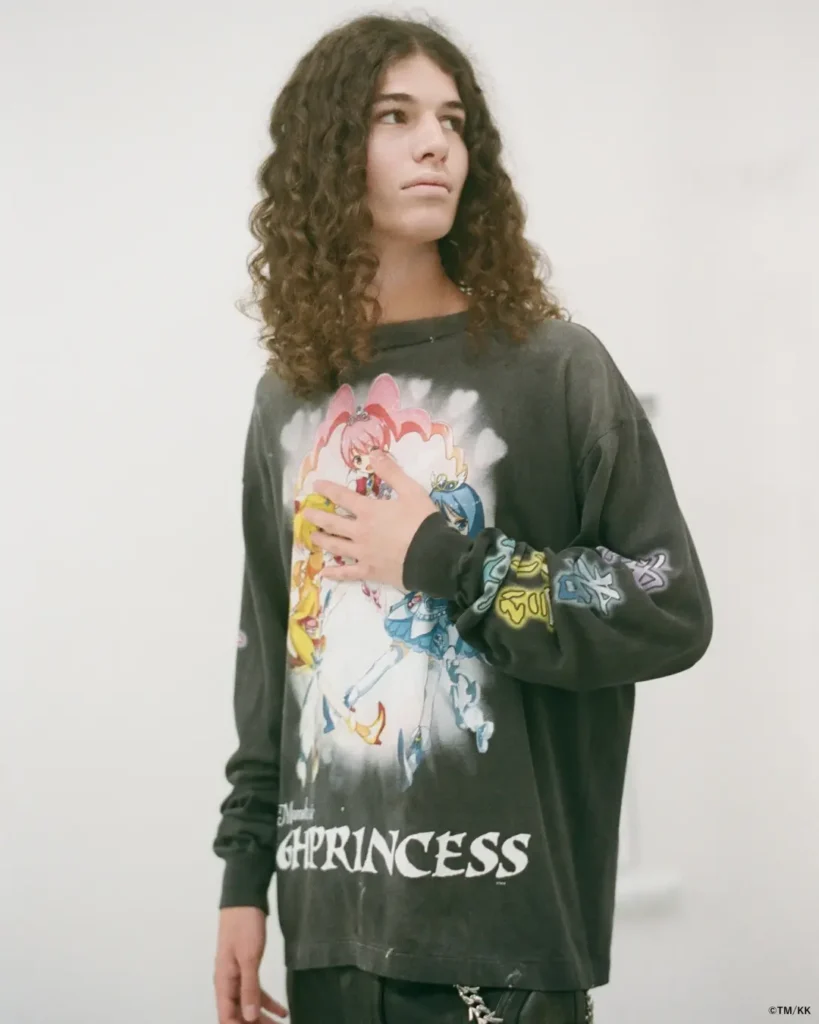 Model with long curly hair wears an oversized black graphic long-sleeve shirt featuring colorful anime-inspired characters and bold typography. The studio portrait is set against a clean white background, highlighting the distressed streetwear aesthetic