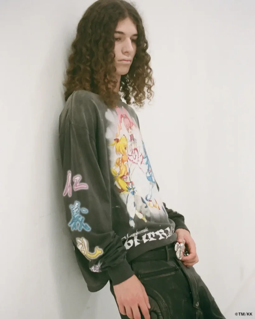 Side-profile fashion shot of a model leaning against a white wall while wearing a faded black long-sleeve graphic shirt. Colorful character artwork and sleeve graphics emphasize the garment’s manga-inspired design language