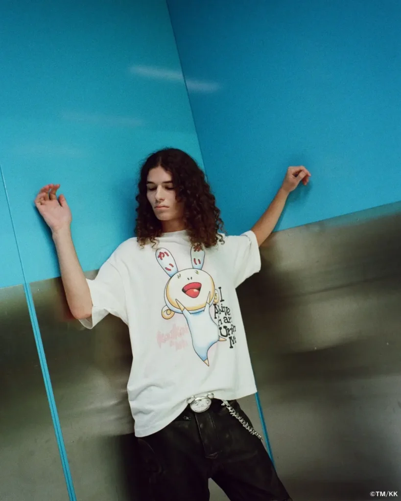Model with shoulder-length curly hair poses against vivid blue wall panels and brushed metal surfaces, wearing an oversized white graphic T-shirt featuring a playful rabbit-like character illustration. Black trousers, a chain wallet, and a relaxed stance give the look a contemporary streetwear edge