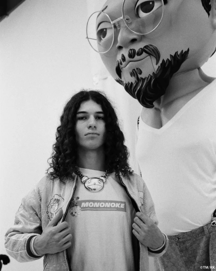 Black-and-white editorial portrait of a model wearing a Mononoke-themed graphic T-shirt, quilted jacket, and oversized character pendant necklace. A large stylized figure sculpture looms beside them, creating a striking contrast between fashion and pop-art influence